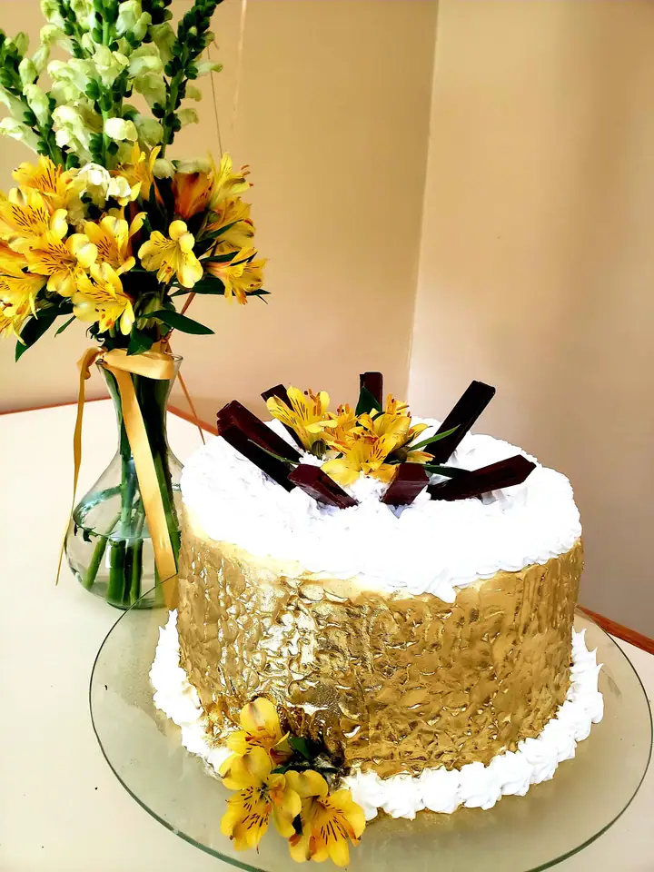 Bolo com lateral dourada, chocolates e flores amarelas no topo