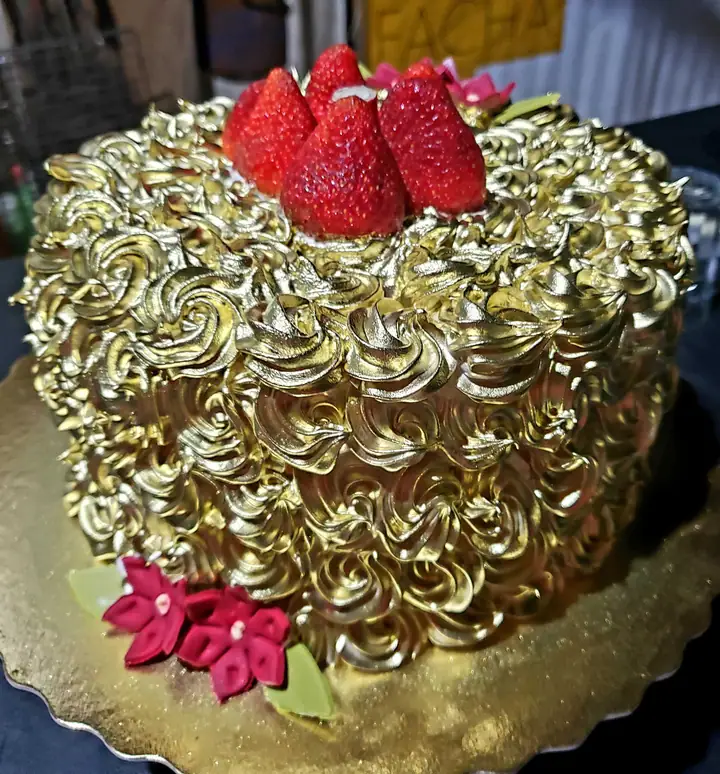 Bolo redondo coberto de rosetas de merengue dourado, com morangos e flores no topo