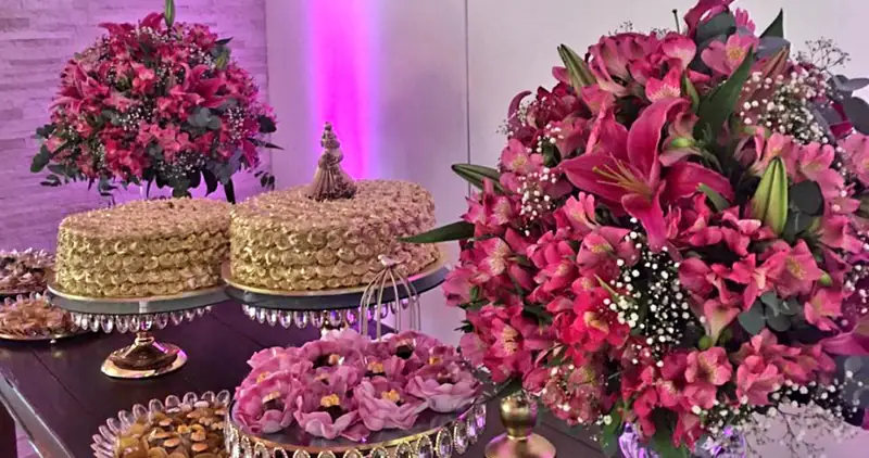 Mesa de festa com dois bolos de merengue dourado em boleiras de cristal, doces e arranjos de flores pink